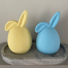 Load image into Gallery viewer, 2 Jesmonite eggs with Bunny ears sat on an oval jesmonite trinket tray. Bunny colours one Yellow, one is blue. Jesmonite Trinket Tray is dark grey marble.