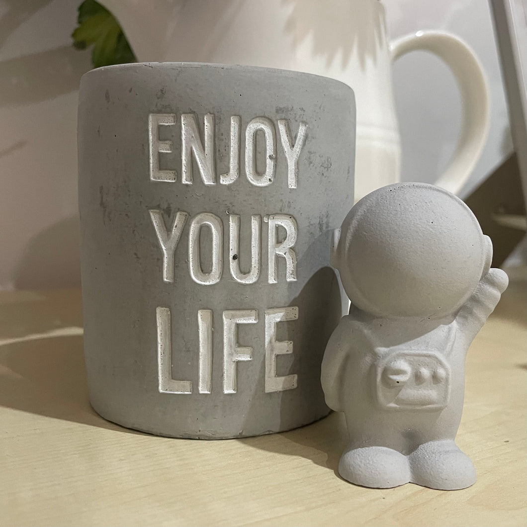 small, grey coloured astronaut waving with one hand up and one hand next to his side, has decorative buttons on tummy and a small pack on his back. this is displayed next to a candle holder saying enjoy your life. 