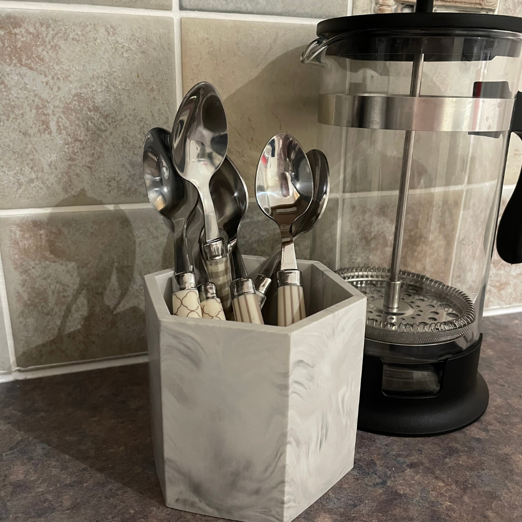 Image shows a grey marble pot from Troody scrumptious made from jesmonite with a smooth finish holding some teaspoons with a coffee pot behind it. 