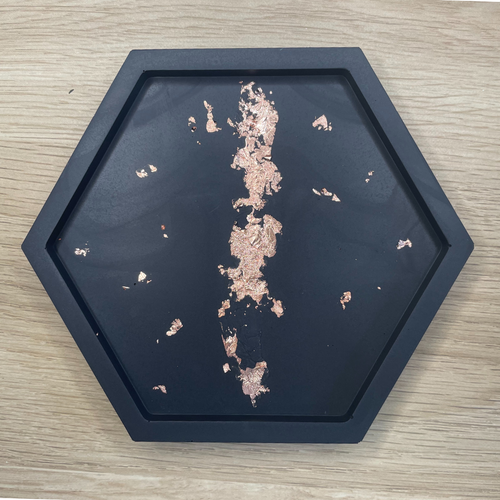 image shows, a hexagon shaped Jesmonite trinket dish or coaster, in a black colour with a copper foil design through the centre as a line and small speckles around the middle of the design. the sides to the dish are low. 